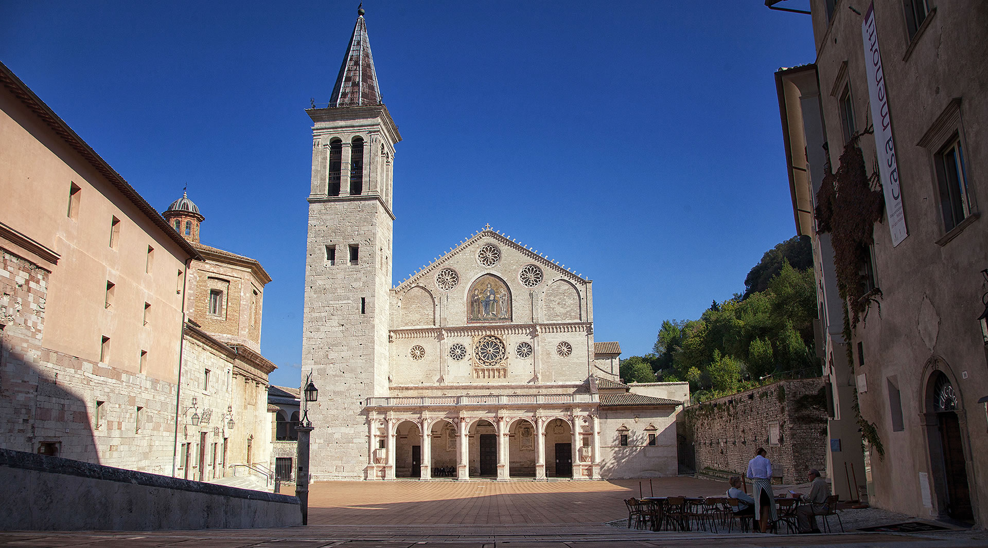 Kathedrale von Spoleto aus weißem und rosafarbenem Stein, mit romanischer Fassade, Rosetten und Portikus, mit Blick auf einen sonnigen Platz.