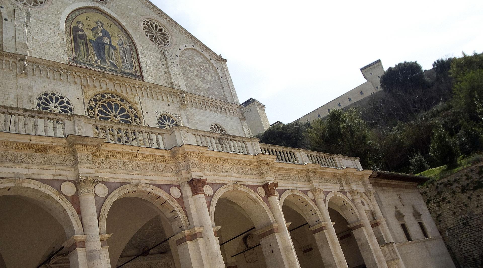 Fassade des Doms von Spoleto mit Bogenportikus, Rosetten, goldenem Mosaik und der Rocca Albornoziana im Hintergrund.