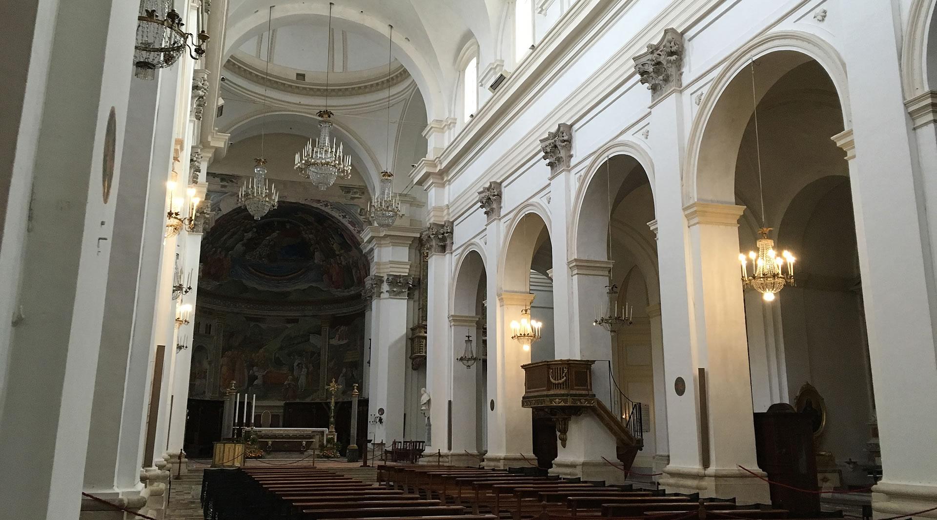 Innenraum des Doms von Spoleto mit weißem Schiff, Kronleuchtern, hölzener Kanzel und Fresken in der Apsis.
