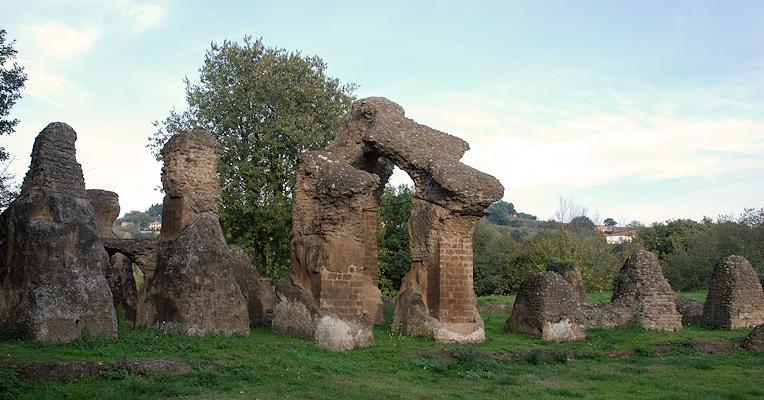 Ruins of the Roman amphitheatre of Ocriculum, with masonry arches surrounded by grass and trees in the background