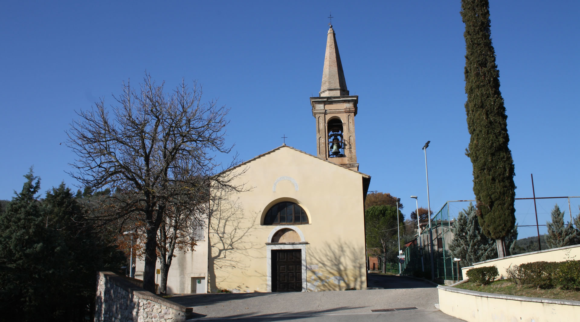 Facciata della chiesa della Madonna del Carmine con campanile a torre, alberi e cielo azzurro sullo sfondo.