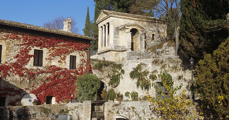 Il Tempietto sul Clitunno in alto e alle spalle di edificio coperto di edera rossa accanto a un canale