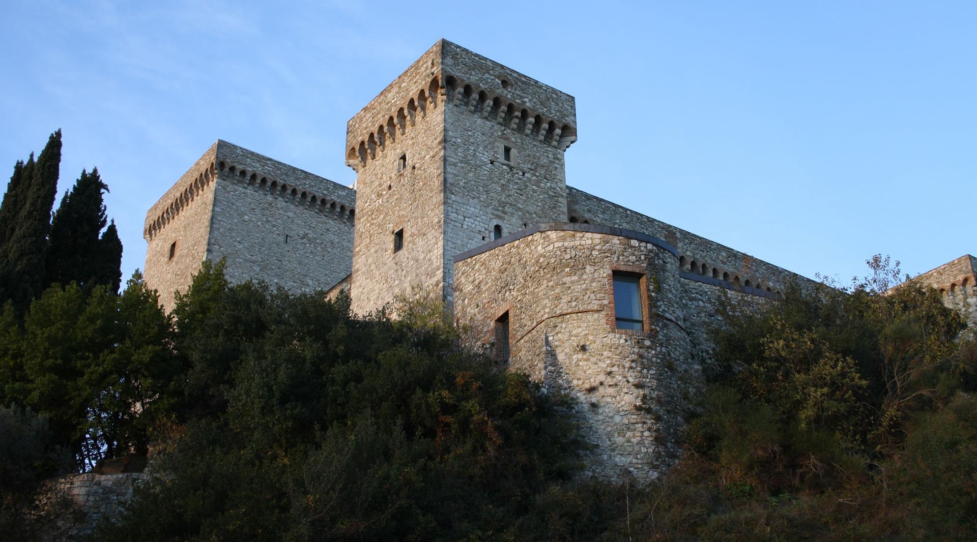 Side view from below of the Fortress of Narni, with its crenellated towers and stone walls rising above the vegetation.