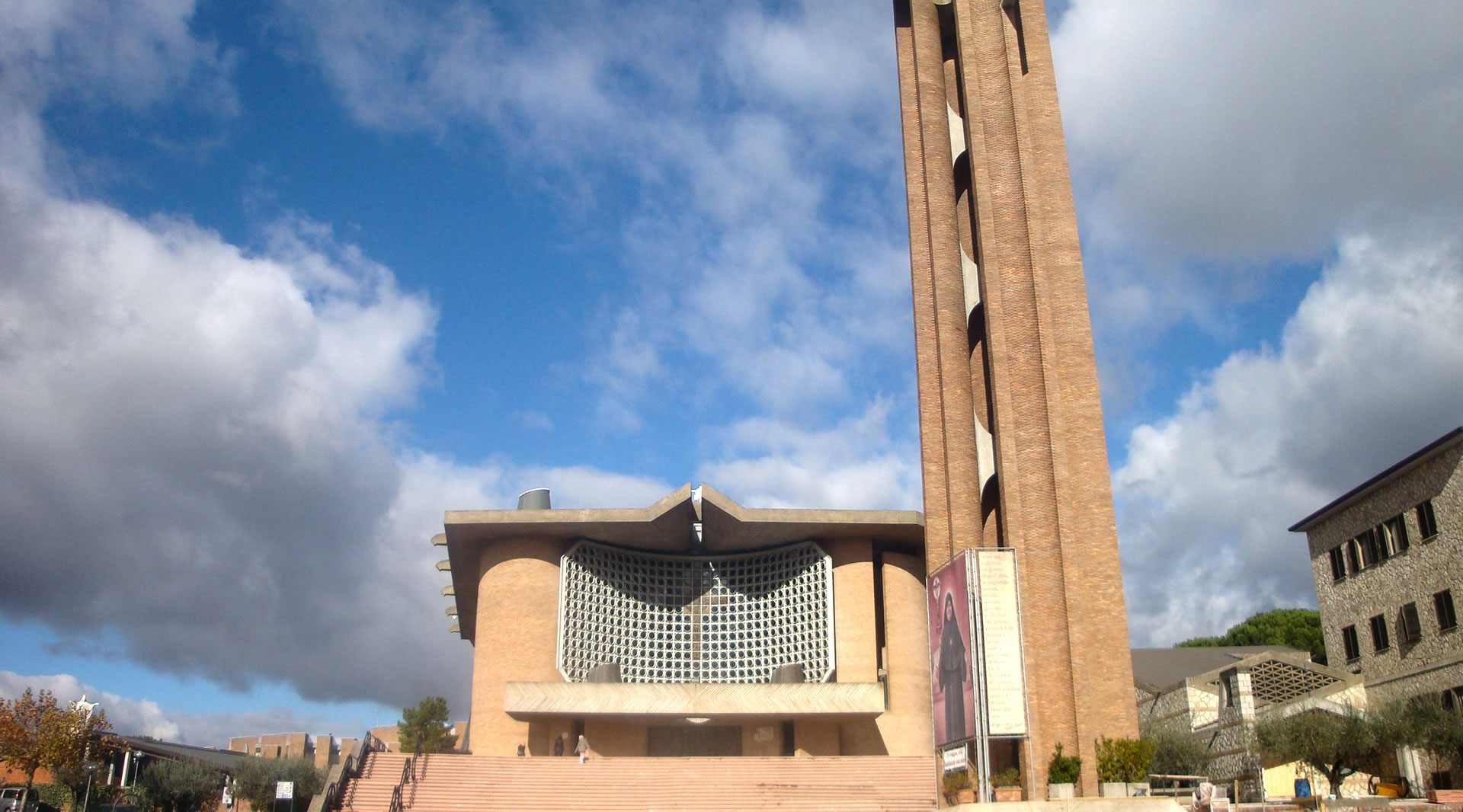 Façade moderne du Sanctuaire de Collevalenza avec un grand clocher en briques, un ciel bleu et nuages en arrière-plan.