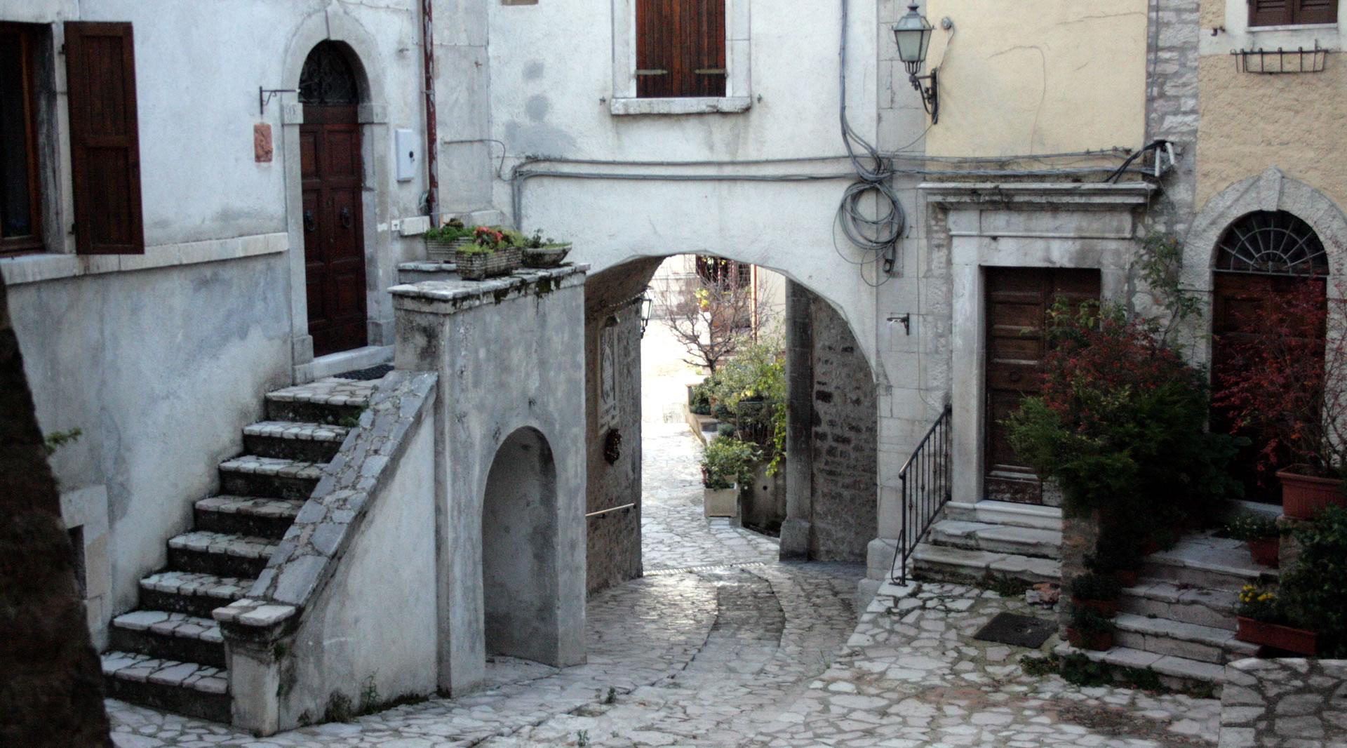 Vicolo in pietra con archi, scalinate e case storiche che si affacciano sulla piazza con pavimento in pietra.
