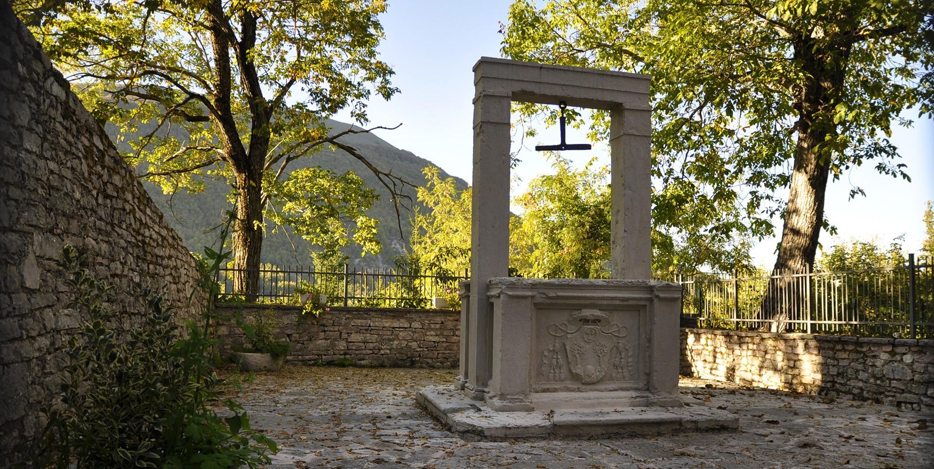 Pozzo storico in pietra con architrave e stemma nobiliare della Famiglia poli, circondato da alberi e mura a Usigni, Poggiodomo.