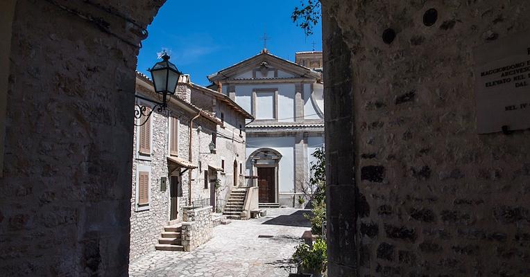 Vista della chiesa di San Salvatore a Usigni attraverso un arco in pietra, con case storiche e strada acciottolata.