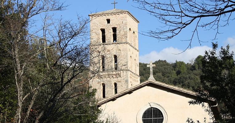 Facade and bell tower of the Abbey of San Pietro in Valle in Ferentillo, with a backdrop of trees and blue sky. The facade features a large circular rose window in the center, while the bell tower, with a quadrangular plan and arched windows, stands behind it.