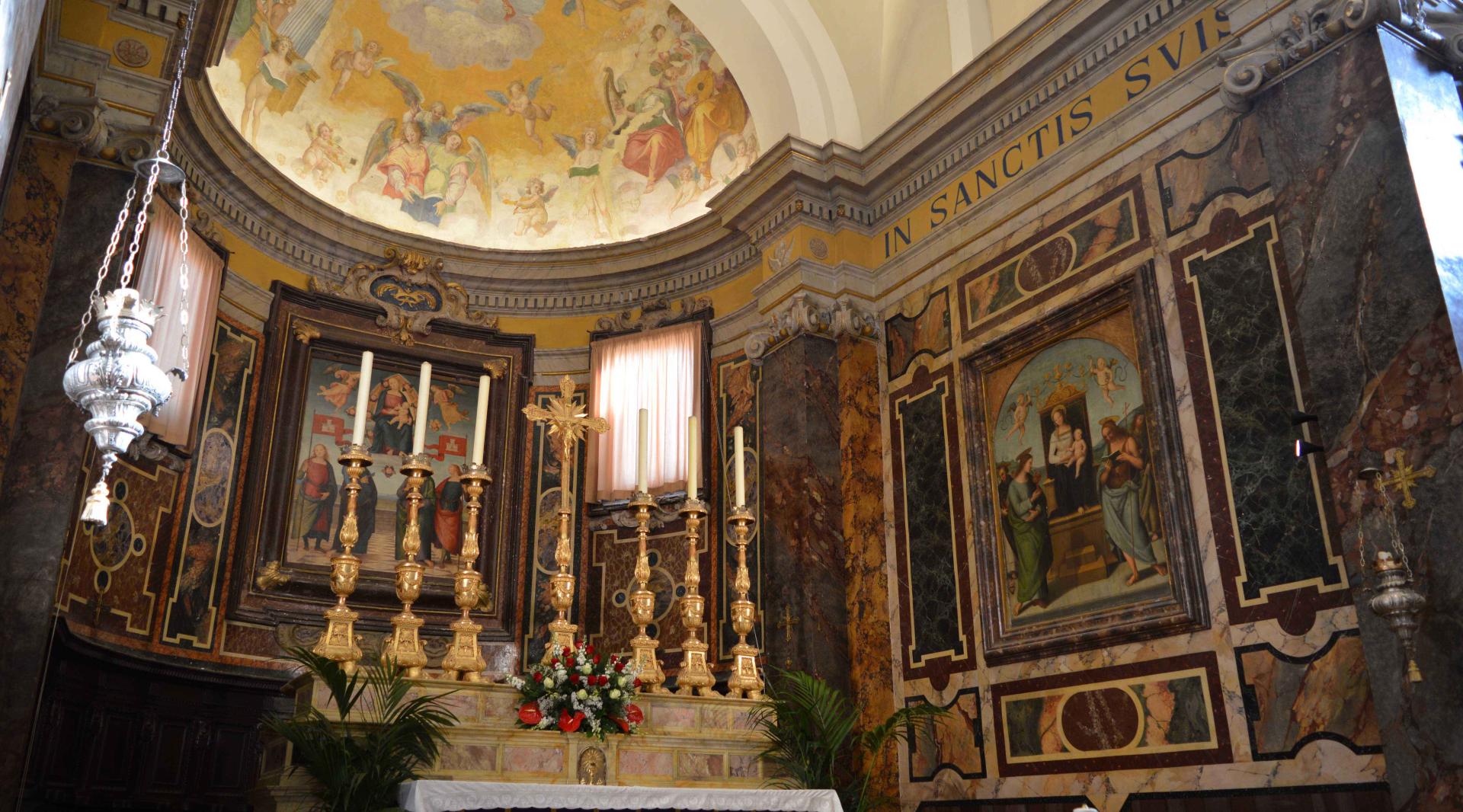 Close-up of the altar adorned with chandeliers, a golden crucifix, and a thurible. In the background of the apse, one can admire a work by Perugino: the "Madonna in Glory between the Holy Protectors Gervasius and Protasius."