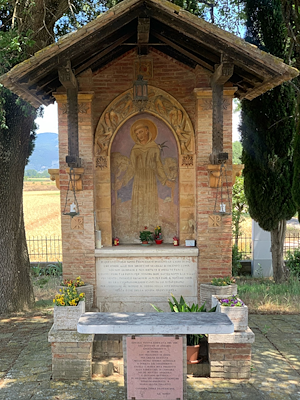 Votive aedicule with a painting of Saint Francis, yellow flowers and red candles, among trees and greenery.