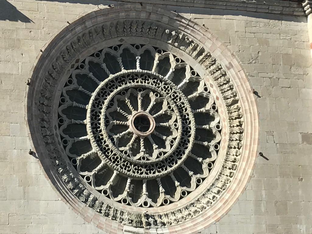 Detail of the Gothic rose window of Todi Cathedral, featuring intricate stone-carved motifs against a backdrop of light-coloured stone walls.