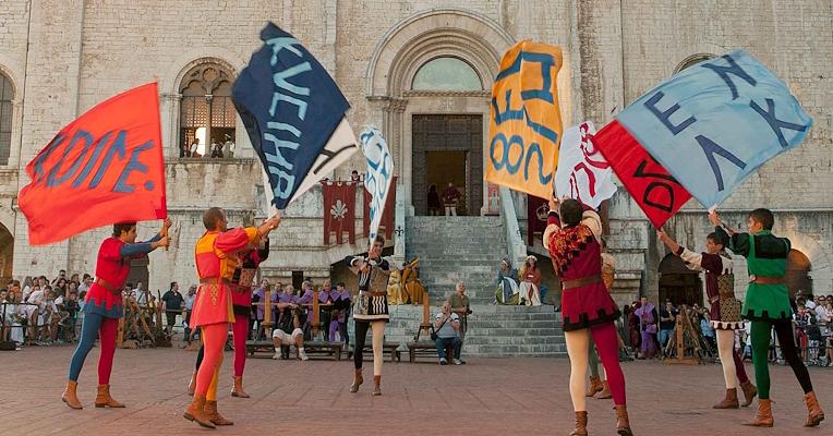 Flag-throwers in historical costume perform in Piazza Grande, Gubbio, waving colourful flags in front of a church.