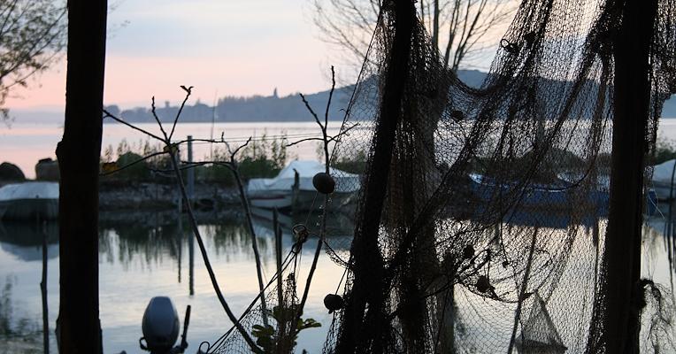 Hanging fishing nets frame Lake Trasimeno at sunset, with moored boats and an island in the background.