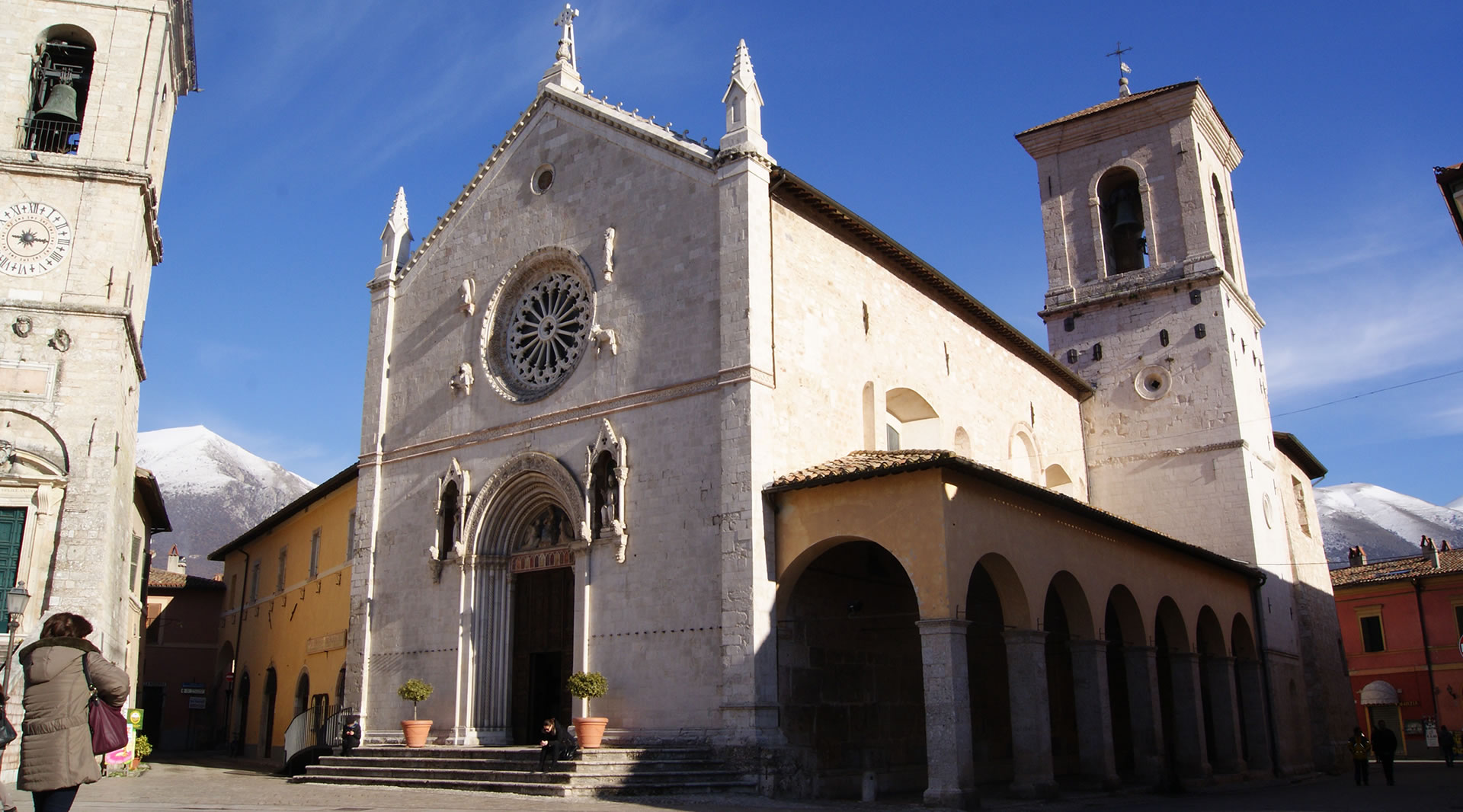 Basilica of San Benedetto in Norcia, with its Gothic façade and rose window, framed by snow-capped mountains under a clear sky.