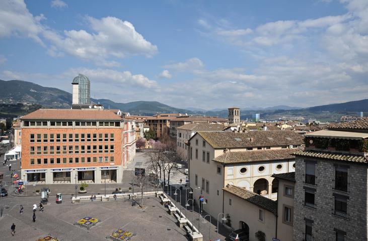 Panorama of Terni’s main square with modern and historic buildings, people walking, and Umbrian hills in the background.
