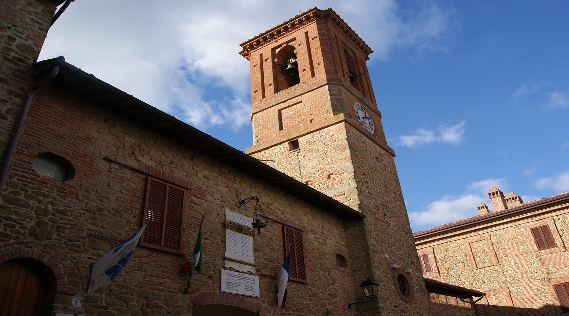 The brick town hall of Paciano, with clock tower and bell tower, under a partly cloudy sky.