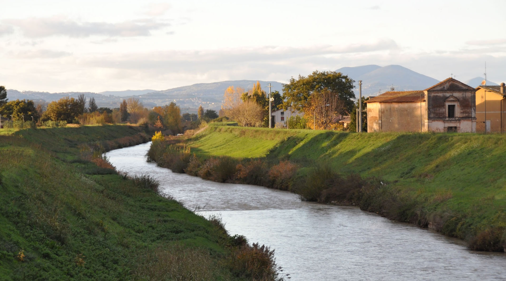 River flowing between Cannara’s green banks, with rural houses and hills lit by the light of the sunset.