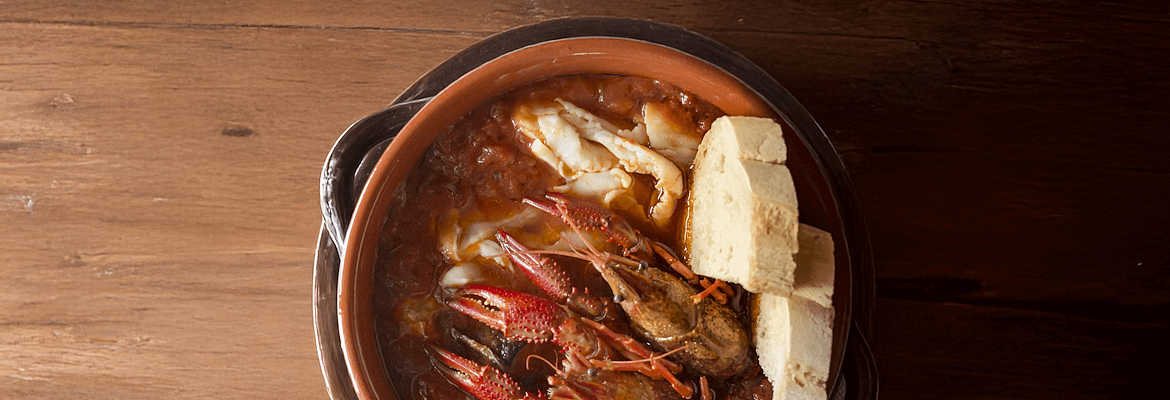 Lake fish stew (tegamaccio) with river crayfish, served in a terracotta pot with slices of bread, on a wooden table.