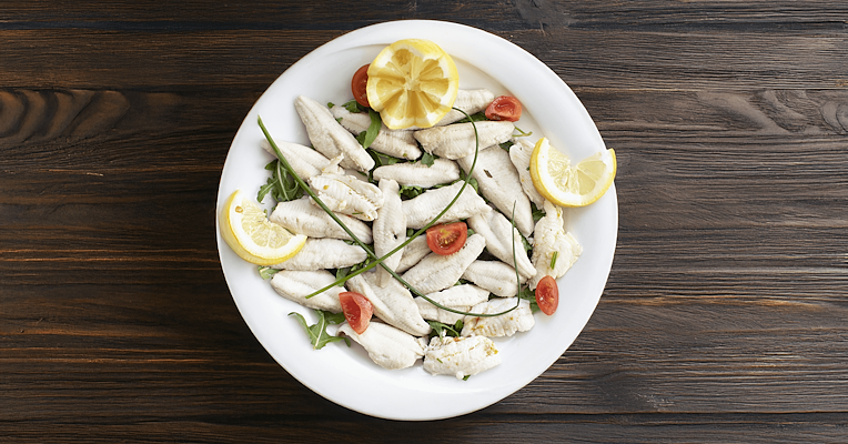 Perch fillets served with rocket, cherry tomatoes, chives, and lemon slices on a white plate, placed on a wooden table.
