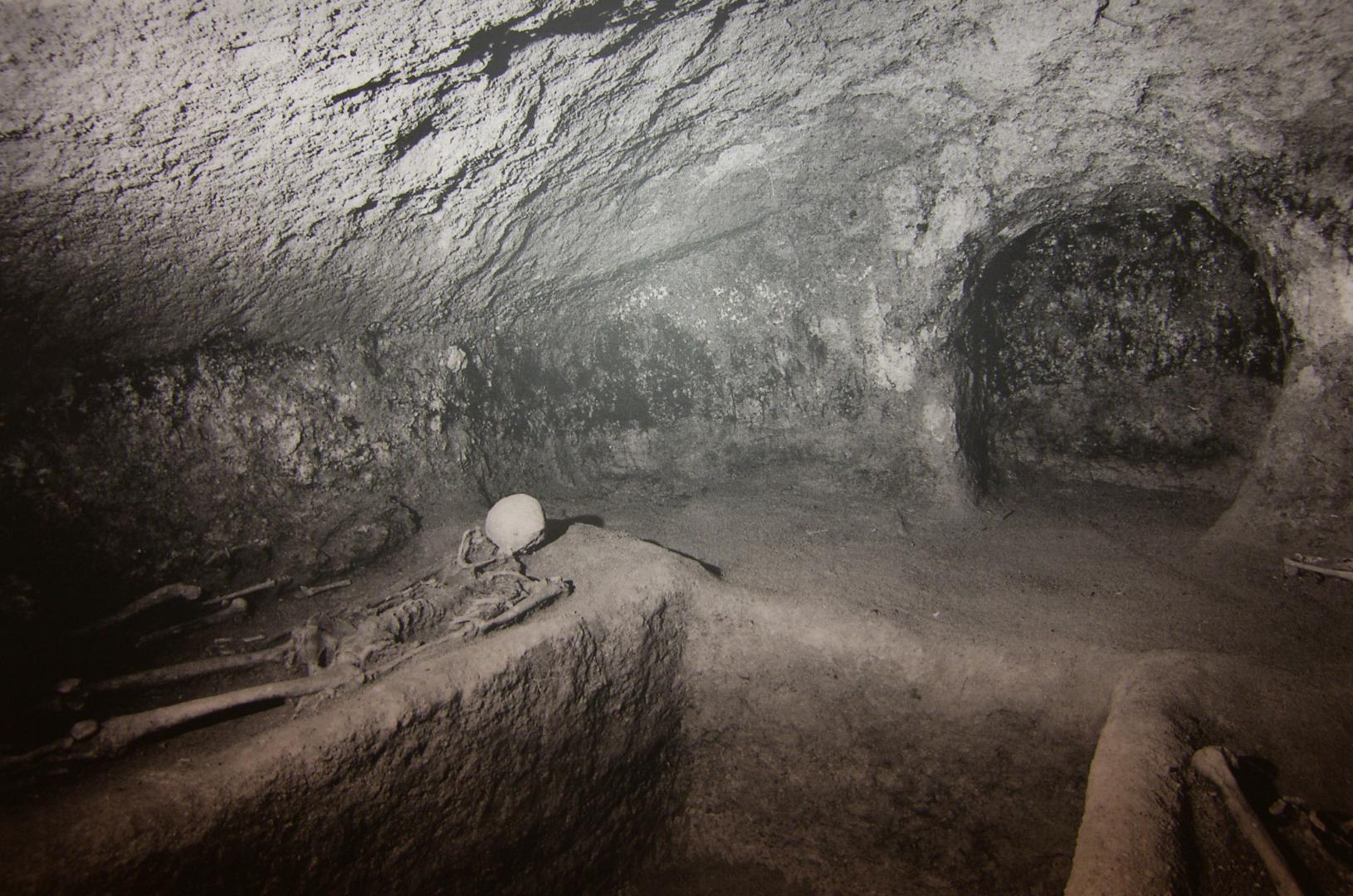 Interior of a rock-cut Etruscan tomb with a human skeleton resting on a platform carved from the rock.
