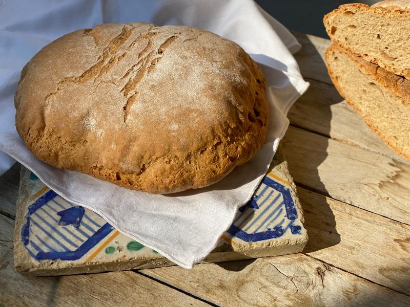 Rustikales Dinkelbrot auf dekorativer Fliese, daneben Brotscheiben, beleuchtet vom natürlichen Licht.