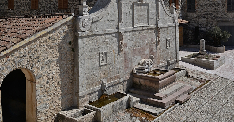 Stone fountain with sculptural decoration and basin in a mountain village, surrounded by ancient buildings and hilly landscape.