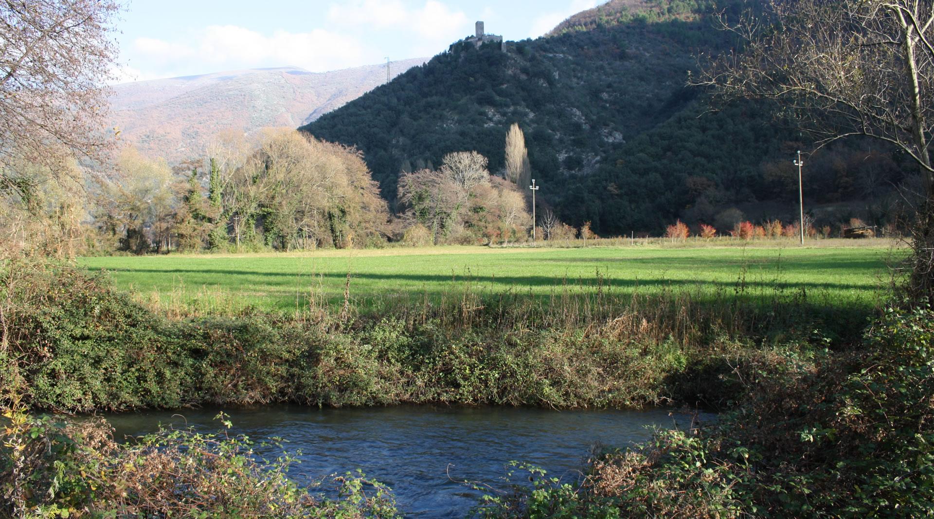 The Nera River flows at the foot of a green valley with woods, fields and a medieval tower overlooking the hill in the background