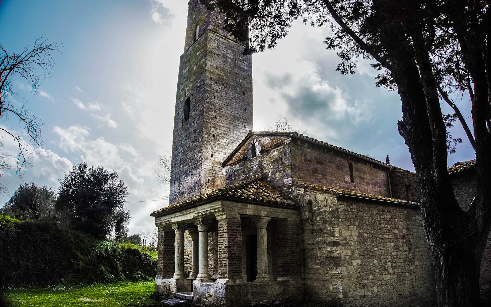 Chiesa di Santa Pudenziana con una torre campanaria in pietra, un portico con colonne e un tetto di tegole. Sullo sfondo, un cielo parzialmente coperto da nuvole e alberi.