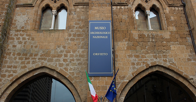 Entrance with flags and a blue banner reading “Museo Archeologico Nazionale – Orvieto” in gold letters