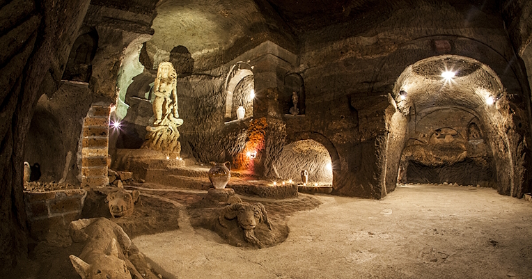 Illuminated underground room with rock-carved statues, arches, and ancient artefacts.