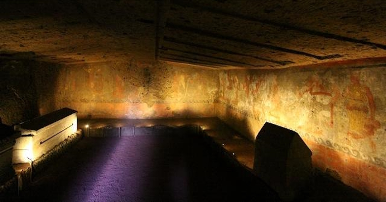 Illuminated underground chamber of an Etruscan tomb with frescoed walls and a raised platform holding cinerary urns