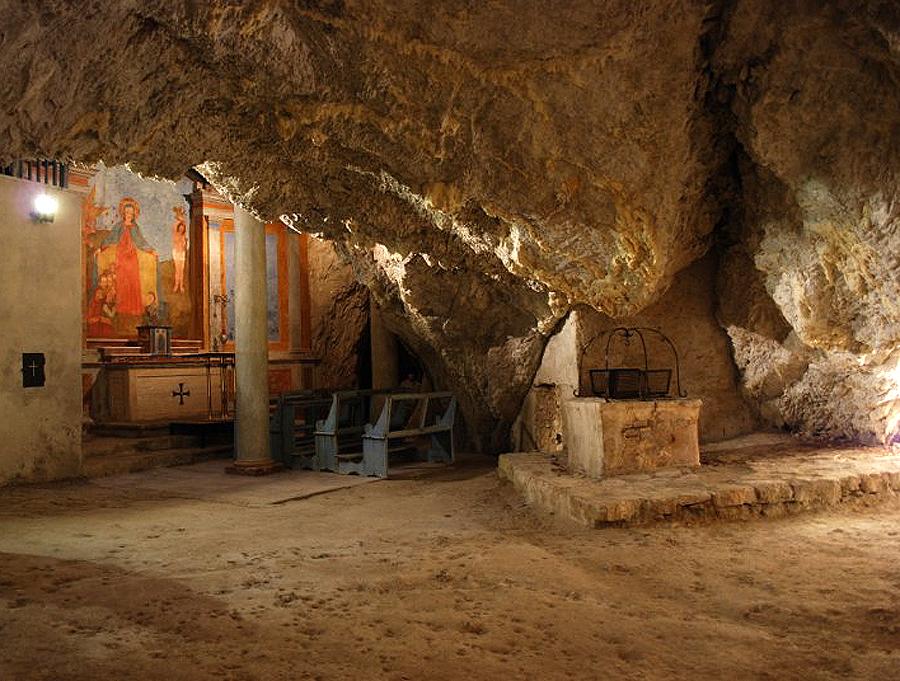 Inneres der Höhle der Einsiedelei Madonna del Riparo mit Freskenaltar, Holzbänken und altem Steinbrunnen.