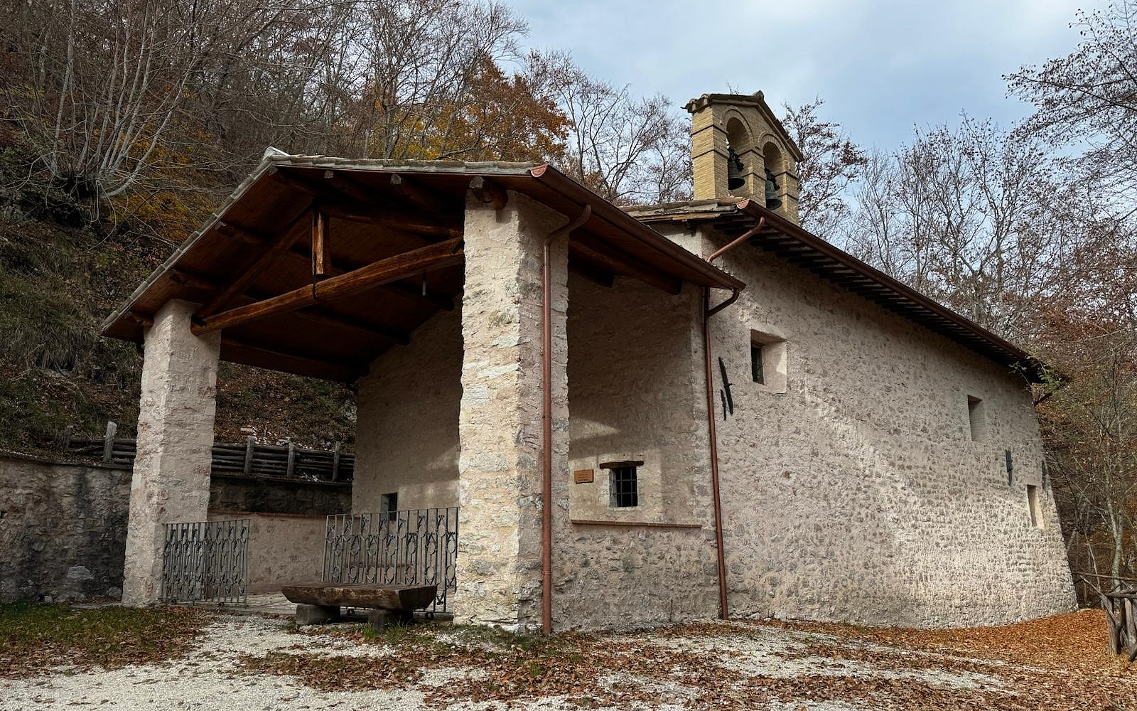 Small stone hermitage with a bell tower and wooden porch, nestled among trees on a mountain slope.