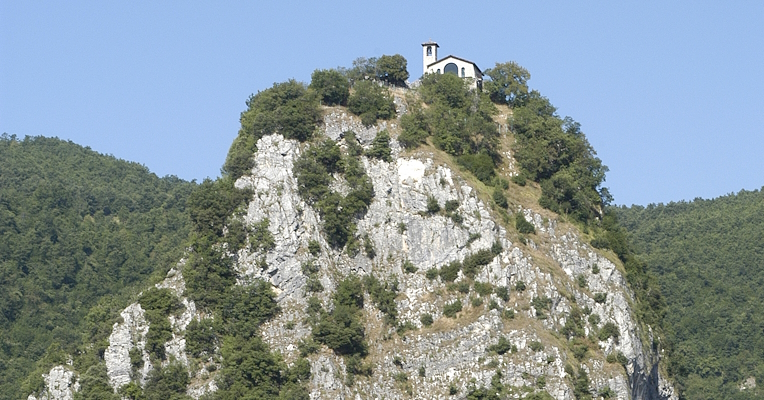 Rocky spur with vegetation and a small church on top, set in a green hilly landscape under a clear sky.