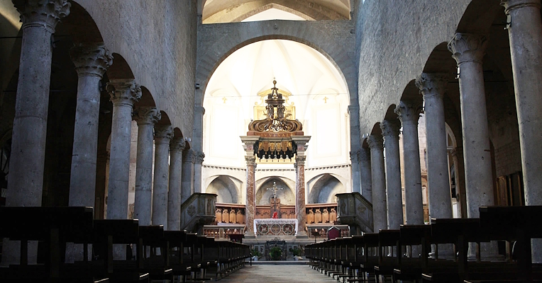 Central nave of the Cathedral of San Giovenale with columns and a vaulted ceiling, a decorated altar at the back, and soft lighting.