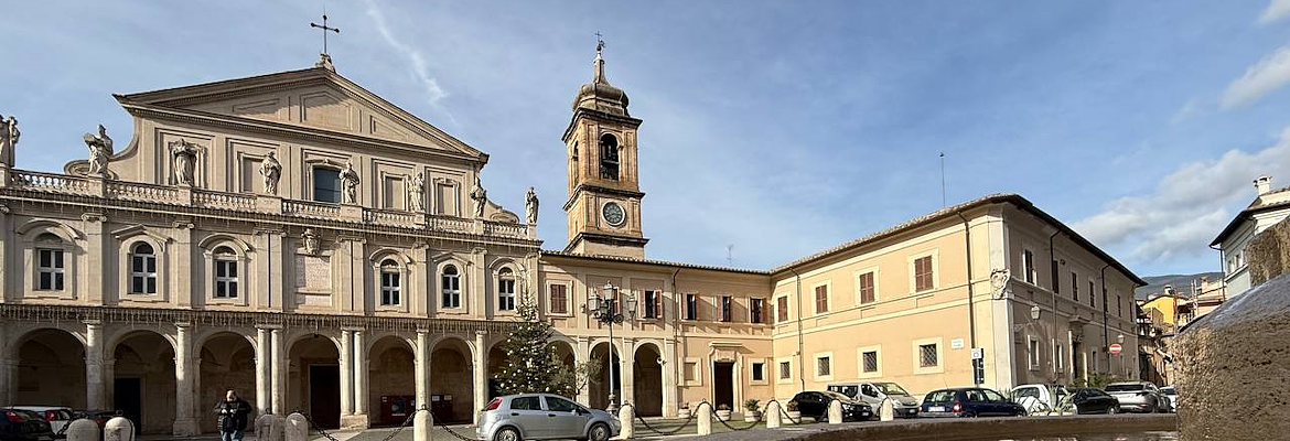 The façade of the Cathedral of Santa Maria Assunta in Terni, with its colonnaded portico and side bell tower, with the fountain in front reflecting the sky and the building adjacent to the cathedral