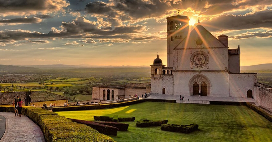 The Basilica of Saint Francis of Assisi at sunset, with the sun filtering through the clouds and illuminating the meadow and valley