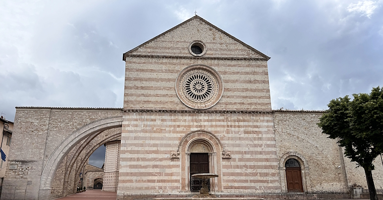 Façade of the basilica of Saint Clare in Assisi, featuring its characteristic pink stone and a fountain in the foreground.
