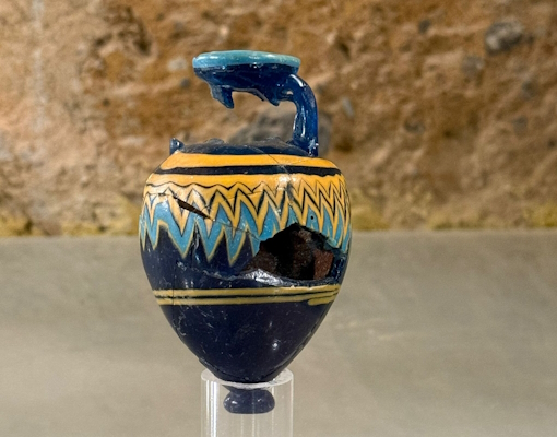 Ancient glass paste unguentarium with fractures on the neck and body, with decorative motifs in blue and yellow on a blue background, displayed on a transparent stand