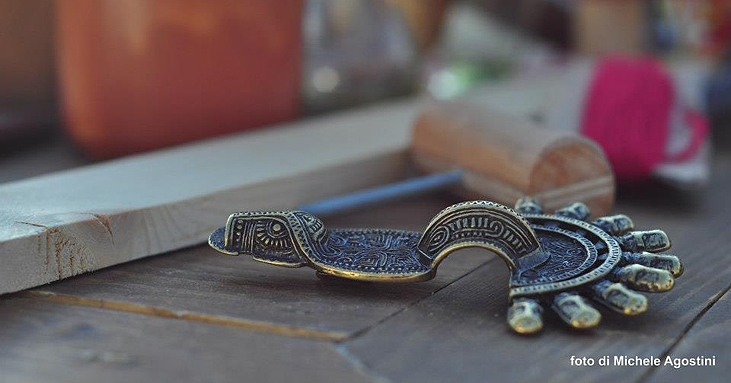 Lombard metal fibula decorated with geometric motifs, resting on a wooden surface, with weaving tools and yarn in the background.
