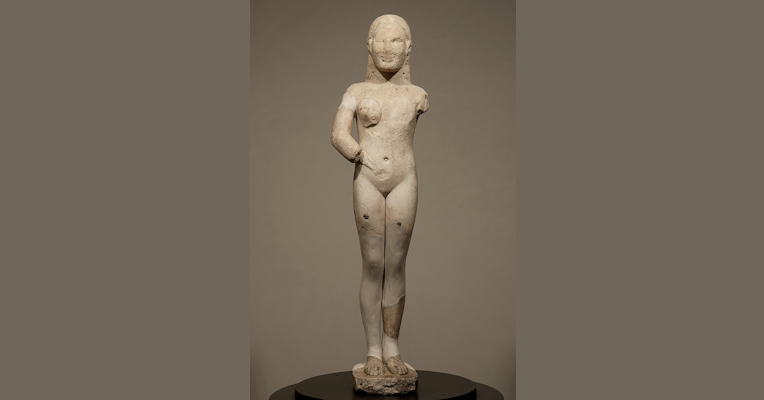 Archaic female statue in white marble, with missing and restored parts, shown in a frontal pose.