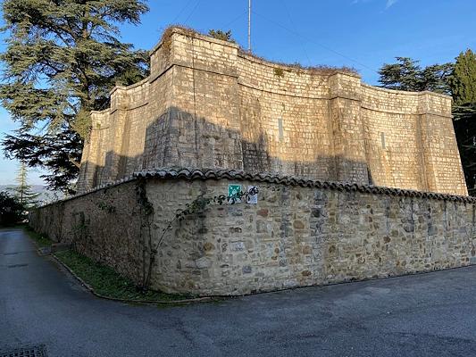 A massive stone structure with fortified walls, surrounded by trees and a lower wall along a paved road.