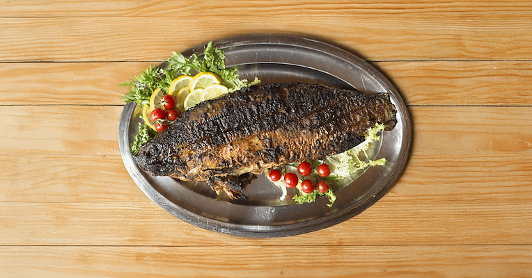 Carp in porchetta on a steel tray, garnished with lemon, salad, and cherry tomatoes on a wooden table.
