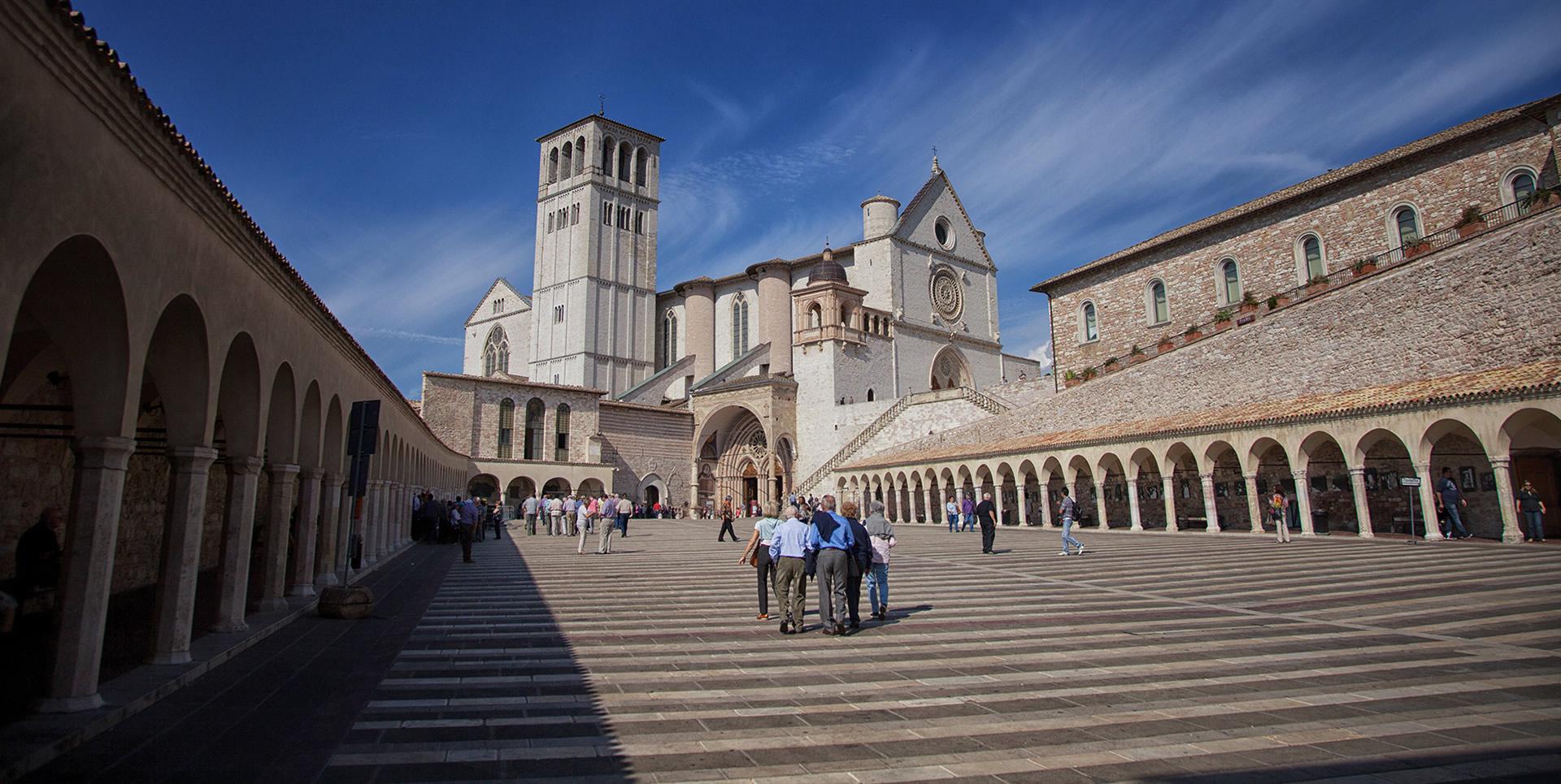 Piazza Inferiore di San Francesco in Assisi, characterised by stone side porticoes and the presence of the Upper Basilica in the background. The central staircase leads to the entrance of the Upper Basilica, highlighting the Gothic architecture of the place.