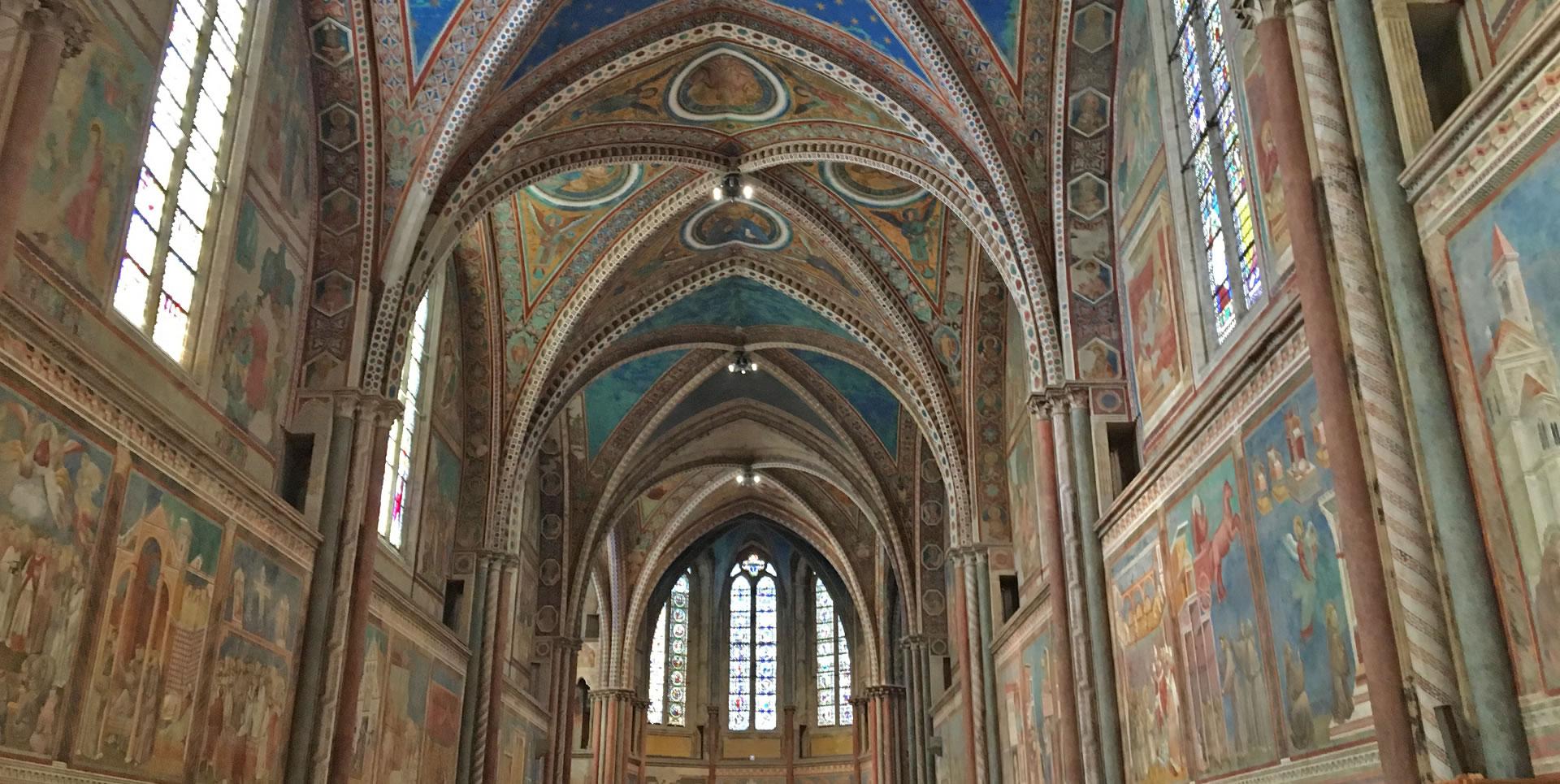 Interior view of the Upper Basilica of St Francis in Assisi, with its high decorated cross vaults. The walls, covered with frescoes by artists such as Giotto and Cimabue, depict scenes from the life of St Francis and biblical stories. Large windows allow natural light to illuminate the interior.