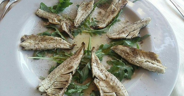 Grilled lake fish fillets arranged in a sunburst on a bed of rocket, seasoned with oil and pepper on a round plate.