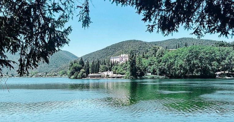 View of Lake Piediluco, with a staircase in the foreground on the left and a small boat moored on the shore to the right; in the background, wooded hills and a pink building nestled among cypress trees.