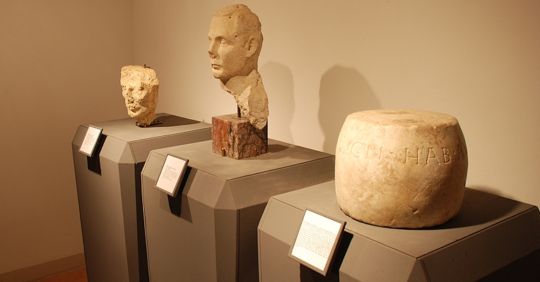 Two stone busts and a cylindrical object with Latin inscription, displayed on pedestals with captions