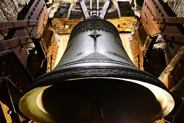Ornate bronze bell seen from below, mounted in a wooden structure inside a bell tower