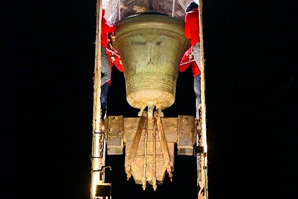 Ornate bronze bell seen from below, mounted in a wooden structure inside a bell tower